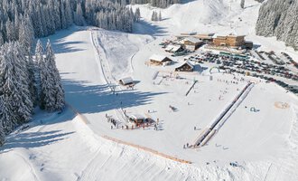 Eine verschneite Landschaft mit Skipisten und einem Restaurant in der Nähe. Hochnebel liegt über den schneebedeckten Bäumen. | © Skischule Hirschegg GmbH