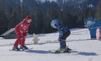 Ein Skilehrer hilft einem Kind beim Skifahren auf einer schneebedeckten Piste. Im Hintergrund sind Bäume und ein blaues Schild mit der Aufschrift "Ski-Kind" sichtbar. | © Skischule Hirschegg GmbH