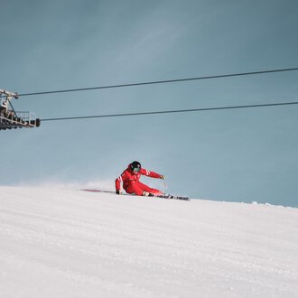 Ein Skifahrer in einem roten Anzug fährt die Piste hinunter. Im Hintergrund ist ein Sessellift vor einem klaren blauen Himmel zu sehen.