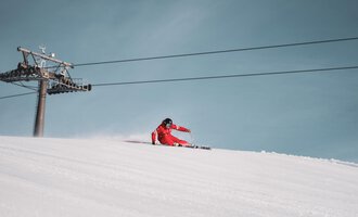 Ein Skifahrer in einem roten Anzug fährt die Piste hinunter. Im Hintergrund ist ein Sessellift vor einem klaren blauen Himmel zu sehen.