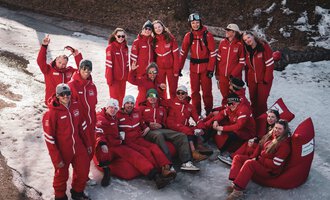 Eine Gruppe von Menschen in roten Overalls posiert fröhlich im Schnee. Sie stehen und sitzen zusammen mit einer entspannenden Atmosphäre.