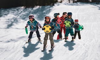 Sechs Kinder in Skiausrüstung stehen auf einem Schneeberg und halten bunte Spielbälle. Die Sonne scheint und die Umgebung ist schneebedeckt.