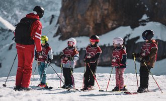 Eine Gruppe von Kindern steht auf Ski im Schnee, während sie von einem Skilehrer instruiert werden. Im Hintergrund sind Berge und ein klarer Himmel zu sehen.