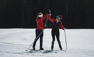 Zwei Frauen in warmer Sportkleidung geben sich beim Langlauf ein High-Five. Sie stehen auf einer schneebedeckten Fläche in einer schönen Winterlandschaft.