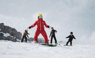Ein Skilehrer in roter Kleidung steht auf einem Snowboard. Hinter ihm üben drei Kinder auf der Piste.