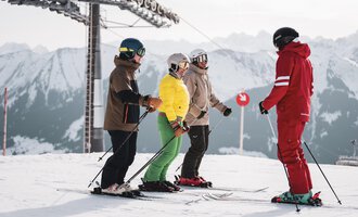 Eine Gruppe von Skifahrern steht auf einer verschneiten Piste. Im Hintergrund sind Berge und ein Skilift zu sehen.