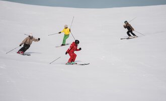 Eine Gruppe von Skifahrerinnen und Skifahrern fährt den Hang hinunter. Sie tragen verschiedene farbige Skibekleidung auf der schneebedeckten Piste.