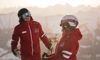 Zwei Skifahrer in roten Jacken unterhalten sich fröhlich in einer verschneiten Berglandschaft. Im Hintergrund sind schneebedeckte Berge und ein klarer Himmel zu sehen.