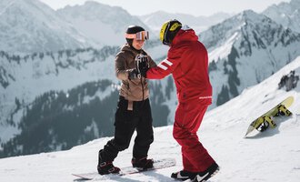Two snowboarders are standing in the snow and helping each other learn. In the background, snow-covered mountains can be seen.