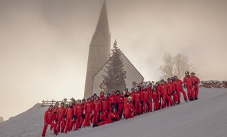 Eine Gruppe von Menschen in roten Skianzügen steht im Schnee vor einer Kirche. Die Landschaft ist winterlich und von Nebel umgeben. | © Skischule Mittelberg | Karl Schuster