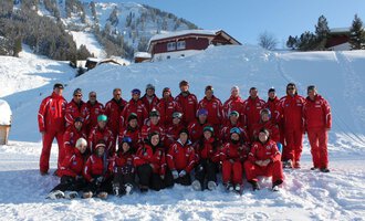 Eine Gruppe von Skifahrern in roten Anzügen posiert im Schnee. Im Hintergrund sind schneebedeckte Berge und Hütten zu sehen. | © Skischule Riezlern GmbH | Frank Felder