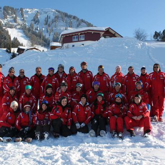 Eine Gruppe von Skifahrern in roten Anzügen posiert im Schnee. Im Hintergrund sind schneebedeckte Berge und Hütten zu sehen. | © Skischule Riezlern GmbH | Frank Felder