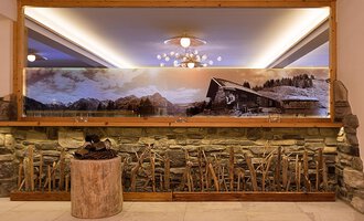 Ein elegantes Interieur mit einer Steinwand und einem großen Spiegel. Im Hintergrund ist ein großflächiges Landschaftsbild mit einer Hütte und Bergen zu sehen. | © Aparthotel Kleinwalsertal Haller GmbH & Co KG