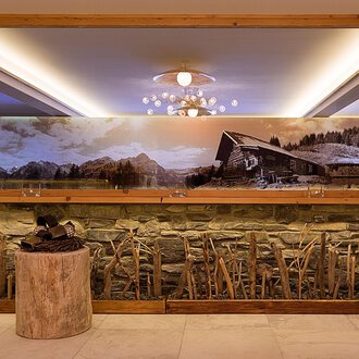 © Aparthotel Kleinwalsertal Haller GmbH & Co KG An elegant interior with a stone wall and a large mirror. In the background, there is a large landscape painting featuring a cabin and mountains. | © Aparthotel Kleinwalsertal Haller GmbH & Co KG