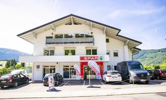 Ein modernes Geschäftshaus mit dem Spar-Logo im Vordergrund. Zahlreiche Autos parken auf dem Parkplatz vor der Tür. | © SPAR