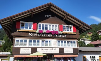 Ein traditionelles Holzhaus mit roten Fensterläden und Blumenschmuck. Der klare Himmel und die umliegenden Berge sorgen für eine idyllische Atmosphäre. | © Sporthaus Edelweiß | N. Lughammer