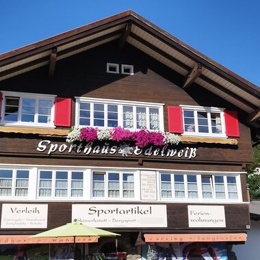 Ein traditionelles Holzhaus mit roten Fensterläden und Blumenschmuck. Der klare Himmel und die umliegenden Berge sorgen für eine idyllische Atmosphäre. | © Sporthaus Edelweiß | N. Lughammer