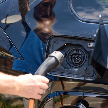 Eine Person lädt ein Elektroauto mit einem Ladekabel auf. Im Hintergrund ist eine moderne Umgebung mit klarem Himmel zu sehen. | © Kleinwalsertal Tourismus | Oliver Farys