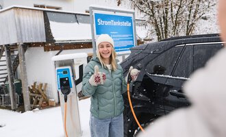 A young woman smiles in front of an electric car charging station in the snow. She gives a thumbs up and is wearing a warm jacket and a hat. | © Kleinwalsertal Tourismus | Oliver Farys