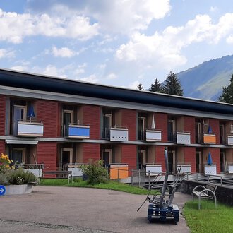 Ein modernes Gebäude mit mehreren Balkonen in einer grünen Umgebung. Im Hintergrund sind Berge und ein blauer Himmel zu sehen. | © Kleinwalsertal Tourismus | Veronika Senn