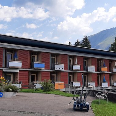 Ein modernes Gebäude mit mehreren Balkonen in einer grünen Umgebung. Im Hintergrund sind Berge und ein blauer Himmel zu sehen. | © Kleinwalsertal Tourismus | Veronika Senn