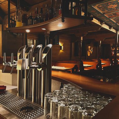 Eine gemütliche Bar mit mehreren Zapfhähnen und einem gut sortierten Getränkeangebot. Im Hintergrund sind Holztische und ein warmes Ambiente zu sehen. | © Taverne | Sead Sulejmani
