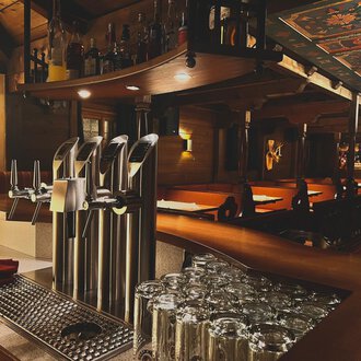 © Taverne | Sead Sulejmani Eine gemütliche Bar mit mehreren Zapfhähnen und einem gut sortierten Getränkeangebot. Im Hintergrund sind Holztische und ein warmes Ambiente zu sehen. | © Taverne | Sead Sulejmani