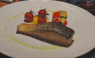 Ein zartes Stück gebratener Fisch liegt auf einem weißen Teller. Daneben sind frische Kirschtomaten und gemischtes Gemüse angerichtet. | © Taverne | Sead Sulejmani