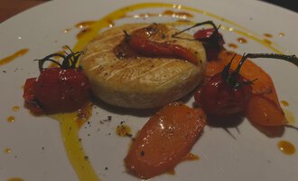 Ein Teller mit gebackenem Käse, umgeben von buntem Gemüse wie Karotten und Kirschtomaten. Der Rand ist mit einer aromatischen Sauce verziert.