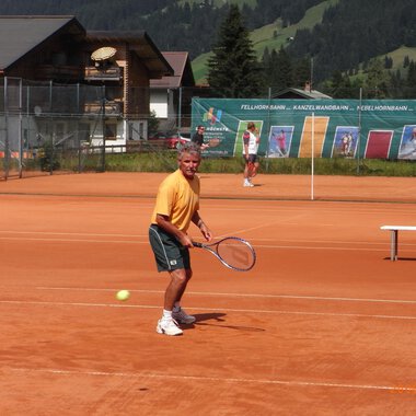 Ein Mann spielt Tennis auf einem roten Platz mit Blick auf die Berge. Im Hintergrund sind weitere Spieler und eine Tenniswand zu sehen. | © Sportverein Kleinwalsertal