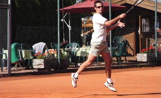Ein Mann spielt Tennis auf einem Sandplatz. Im Hintergrund sind Stühle und Tische sowie ein paar Pflanzen zu sehen. | © Sportverein Kleinwalsertal
