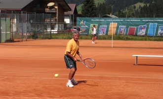 Ein Mann spielt Tennis auf einem roten Platz mit Blick auf die Berge. Im Hintergrund sind weitere Spieler und eine Tenniswand zu sehen. | © Sportverein Casino Kleinwalsertal