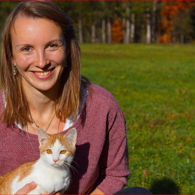 A smiling woman is sitting on a meadow holding an orange-and-white cat. Autumn trees can be seen in the background. | © Tierarztpraxis Oberstdorf | Regina Baumgartner