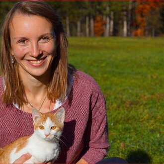 Eine lächelnde Frau sitzt auf einer Wiese und hält eine orange-weiße Katze. Im Hintergrund sind herbstliche Bäume zu sehen. | © Tierarztpraxis Oberstdorf | Regina Baumgartner