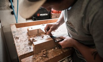 A craftsman is working with a tool on a piece of wood. Wood shavings are lying on the workbench as he makes precise cuts. | © Türtscher Die Schreinermanufaktur