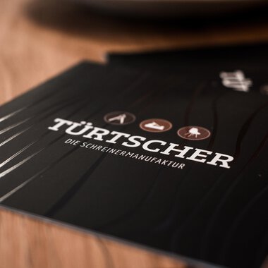 Ein elegantes Menüheft mit der Aufschrift „TÜRTSCHER – die Schreiner-Manufaktur“. Es zeigt verschiedene Holzsymbole auf einem dunklen Hintergrund. | © Türtscher Die Schreinermanufaktur