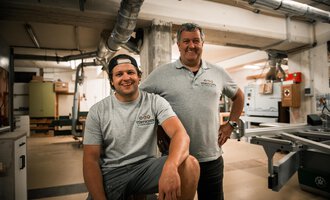 Zwei Männer stehen in einer Werkstatt. Der jüngere Mann sitzt auf einem Hocker, während der ältere Mann stolz neben ihm steht. | © Türtscher Die Schreinermanufaktur