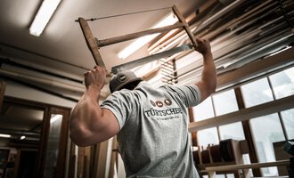 Ein Handwerker hält einen Stuhl über seinen Kopf und arbeitet in einer Werkstatt. Im Hintergrund sind Holzmaterialien und Werkzeuge zu sehen. | © Türtscher Die Schreinermanufaktur