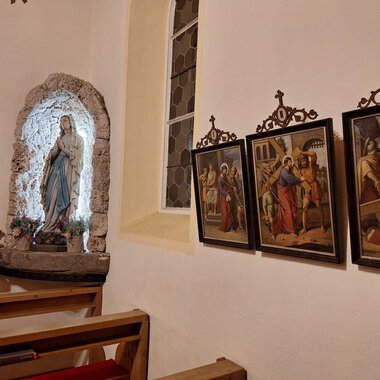 Eine Kirchengalerie mit einer Statue von Maria und mehreren historischen Gemälden an der Wand. Die Atmosphäre ist friedlich und andächtig. | © Unterwestegg-Kapelle Maria Hilf | Tanja Ritsch