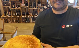 Ein Mann hält einen großen Kuchen in den Händen. Im Hintergrund sind Weinflaschen in Regalen sichtbar. | © Uusziit | Chantal Kessler