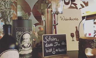 Eine einladende Restaurant-Szene mit Weinkarten, dekorativen Flaschen und einer kleinen Tafel mit der Aufschrift „Schön, dass Du da bist!“. Im Hintergrund sind stilvolle Holzelemente und Kerzen zu sehen. | © Uusziit | Chantal Kessler