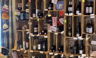 Eine Wand aus Holzregalen mit verschiedenen Weinflaschen und Accessoires. Die Regale sind dekorativ und einige Artikel sind mit Etiketten versehen. | © Uusziit | Chantal Kessler