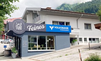 Ein modernes Gebäude mit einem Geschäftsschild „Feines“ und der Volksbank Vorarlberg. Im Hintergrund sind grüne Hügel und ein klarer Himmel zu sehen. | © Volksbank Vorarlberg e. Gen.