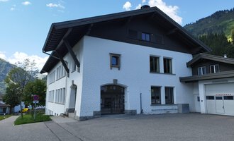 A modern, white building with a large roof. It is located in a mountainous area with green grass and trees. | © Kleinwalsertal Tourismus | N. Lughammer