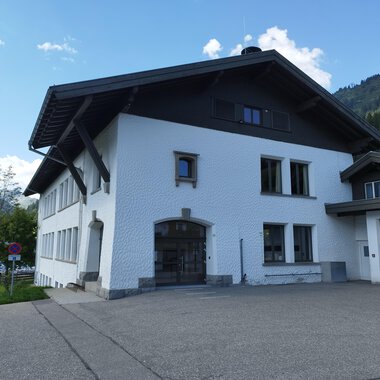 Ein modernes, weißes Gebäude mit einem großen Dach. Es befindet sich in einer bergigen Umgebung mit grünem Gras und Bäumen. | © Kleinwalsertal Tourismus | N. Lughammer