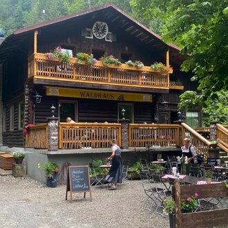 Ein gemütliches Chalet inmitten von Bäumen. Auf der Terrasse sitzen Gäste und genießen die Natur. | © Waldhaus | Yvonne Grosse