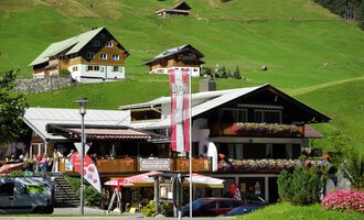 Ein schönes alpenländisches Gebäude mit einer Terrasse und bunten Blumen. Im Hintergrund sind grüne Wiesen und weitere traditionelle Häuser zu sehen. | © Cafe Baad Grund | Roman Schuster