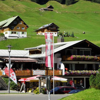 Ein schönes alpenländisches Gebäude mit einer Terrasse und bunten Blumen. Im Hintergrund sind grüne Wiesen und weitere traditionelle Häuser zu sehen. | © Cafe Baad Grund | Roman Schuster