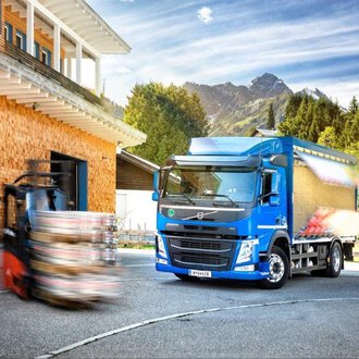 © NUFA | Stefan Kothner A modern truck stands in front of a wooden building with mountains in the background. A forklift is moving nearby while the sun is shining. | © NUFA | Stefan Kothner