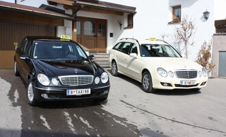 Zwei Taxis stehen vor einem Wohnhaus. Das schwarze Taxi und das weiße Kombi-Taxi sind bereit für Fahrgäste. | © Taxi Beranek | Elmar Beranek
